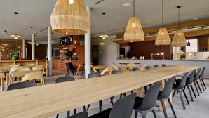 Een ruime eetruimte met lange houten tafels en zwarte stoelen, met geweven hanglampen en een bar in het midden.
