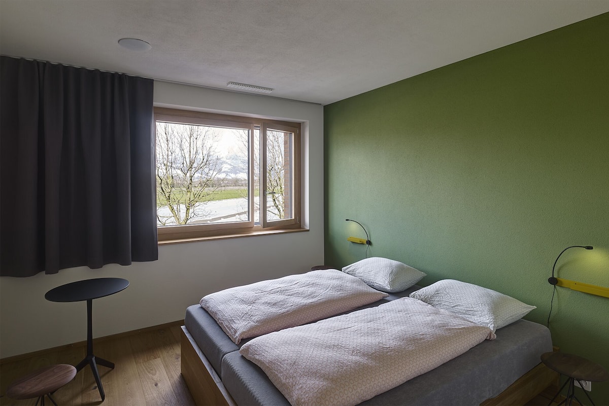 Een slaapkamer met een bed, een klein rond tafeltje, een stoel, een raam met gordijnen en een groene wand.