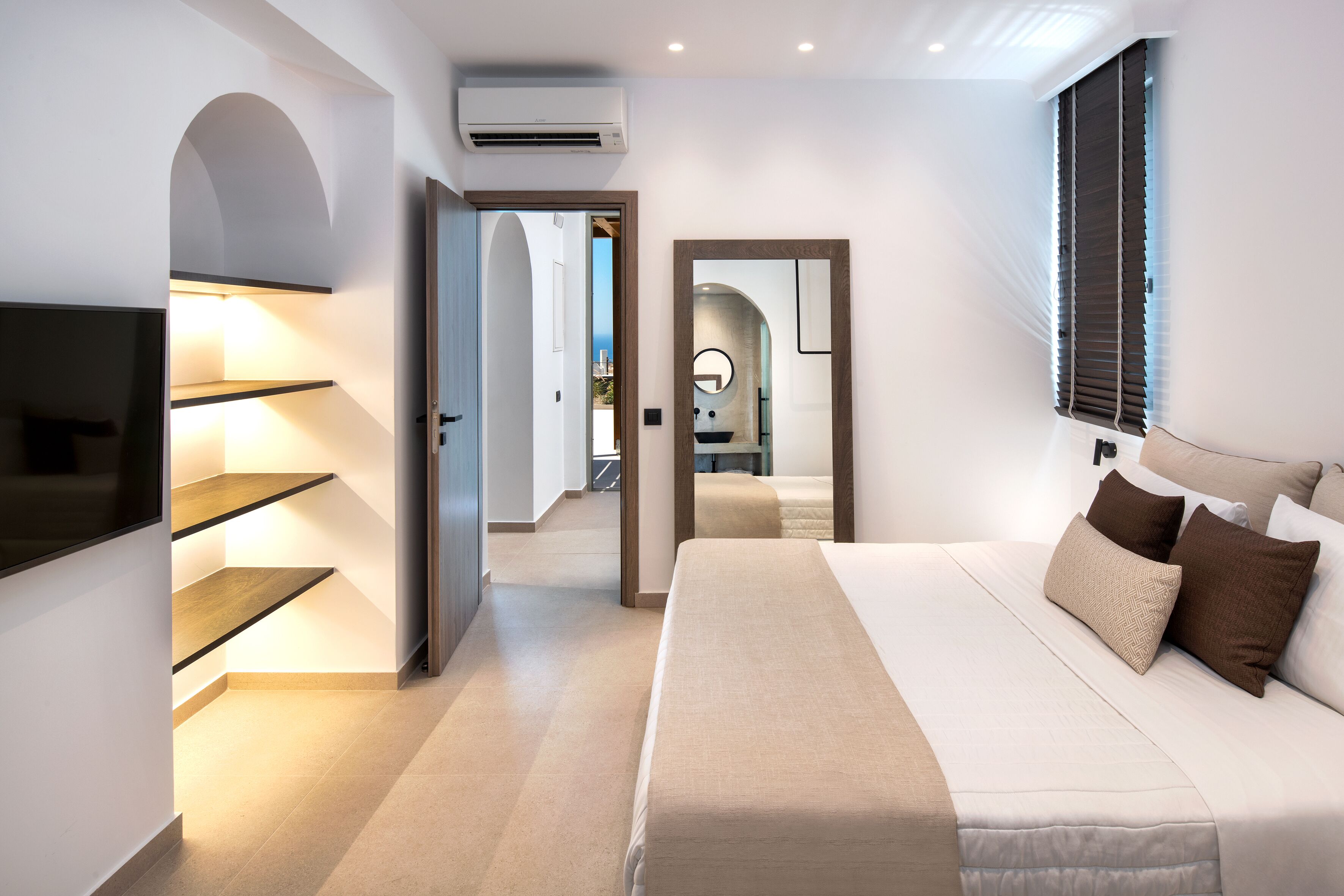 A modern bedroom with a large bed, built-in shelves, a TV, and a view of the sea through the window.