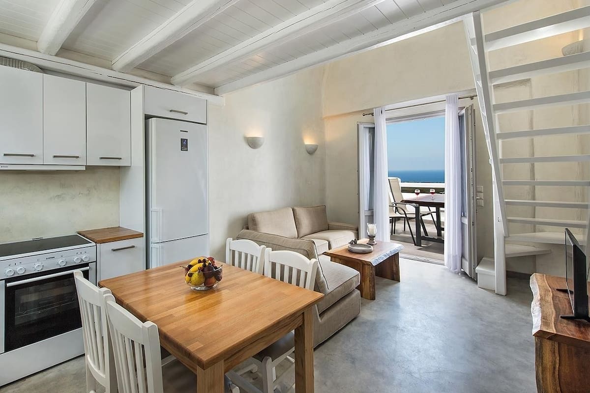 Una cocina compacta con armarios blancos, un refrigerador y una estufa. Hay un comedor de madera con sillas y un pequeño sofá en la sala. Se ve un balcón con vista al mar a través de una puerta abierta.