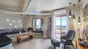 Ein modernes Wohnzimmer mit einer gebogenen Couch, einem Holztisch und einem grauen Sessel. Ein großes Fenster bietet Blick auf das Meer.
