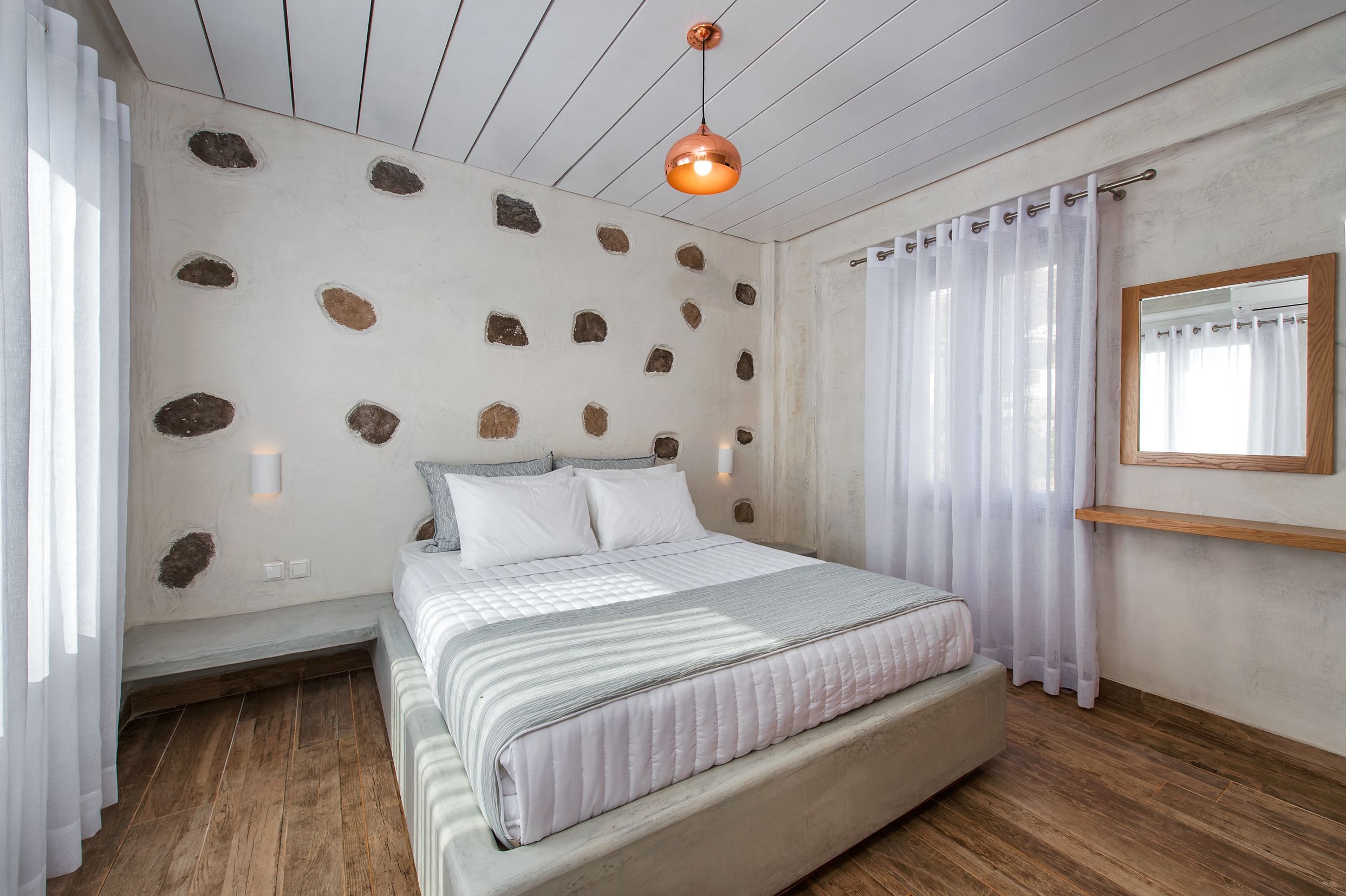 Une chambre à coucher avec un lit, un plancher en bois, des rideaux blancs, un luminaire au plafond et un mur orné de motifs évoquant la pierre.