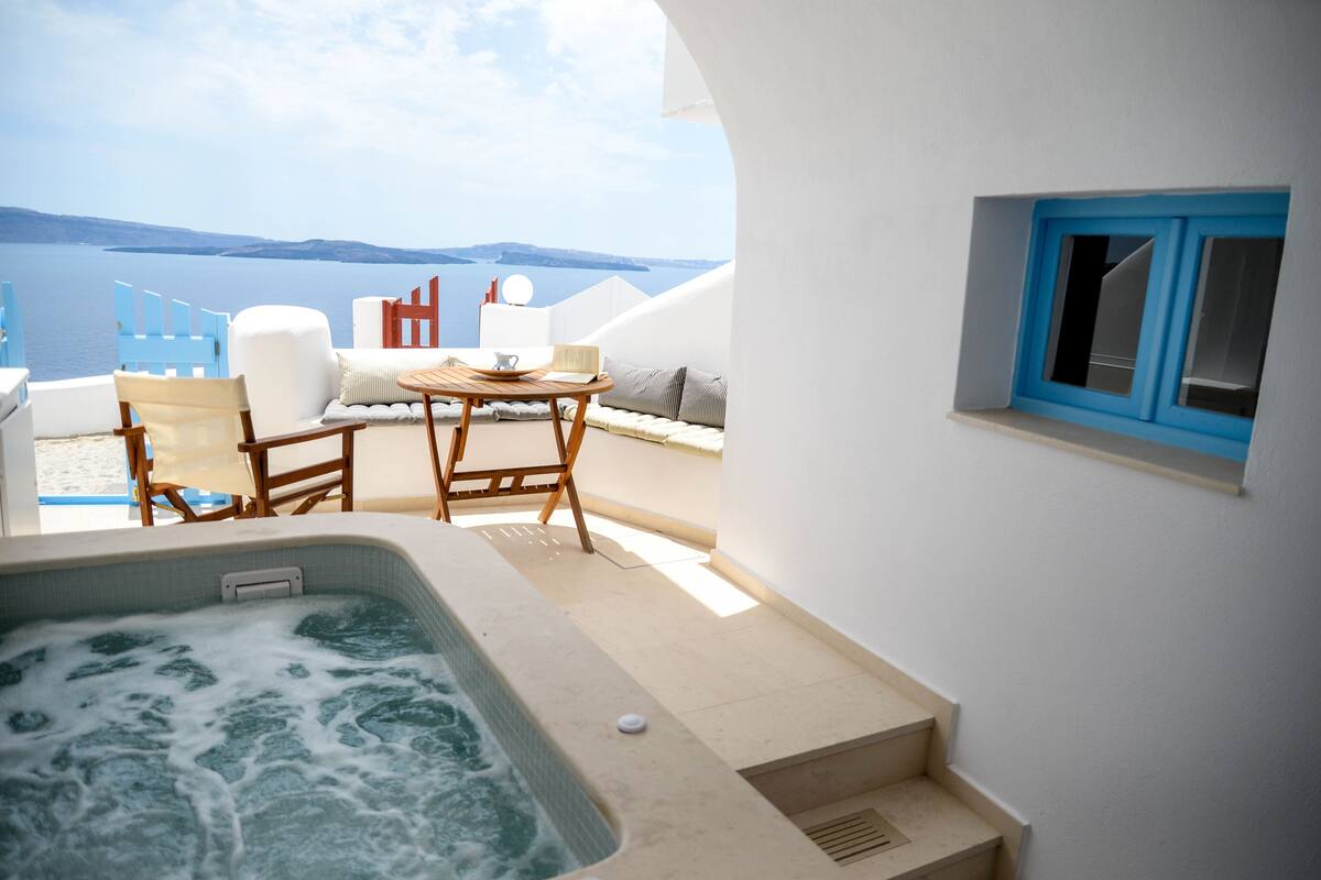 A hot tub on a balcony with a view of the sea, a table with chairs, and a small window.
