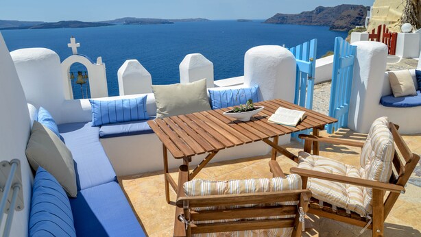 A sunlit outdoor seating area with a wooden table and chairs, a bowl of fruit, and a view of the sea and distant islands. - Genesis Cave Houses (Santorini)