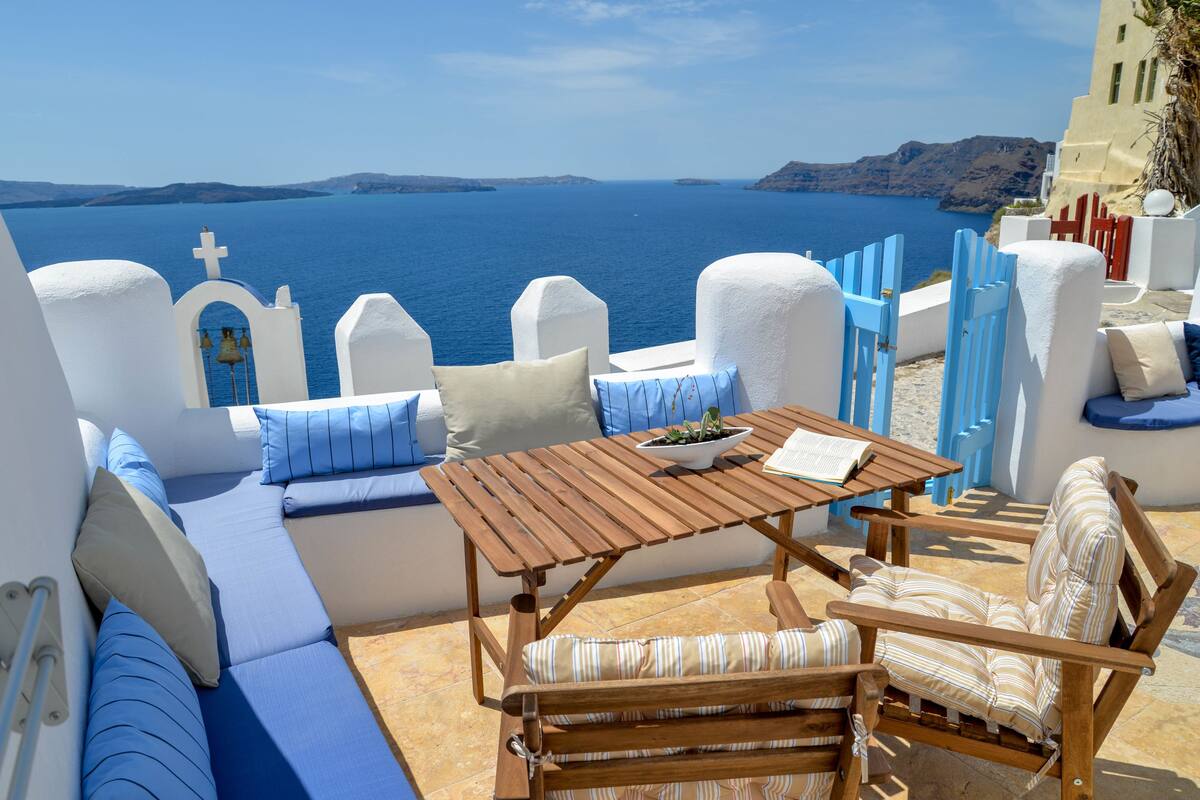 A sunlit outdoor seating area with a wooden table and chairs, a bowl of fruit, and a view of the sea and distant islands.