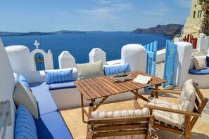 A sunlit outdoor seating area with a wooden table and chairs, a bowl of fruit, and a view of the sea and distant islands. - Genesis Cave Houses (Santorini)