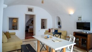 A spacious living area with a white ceiling, a dining table, a sofa, and a television. - Genesis Cave Houses (Santorini)