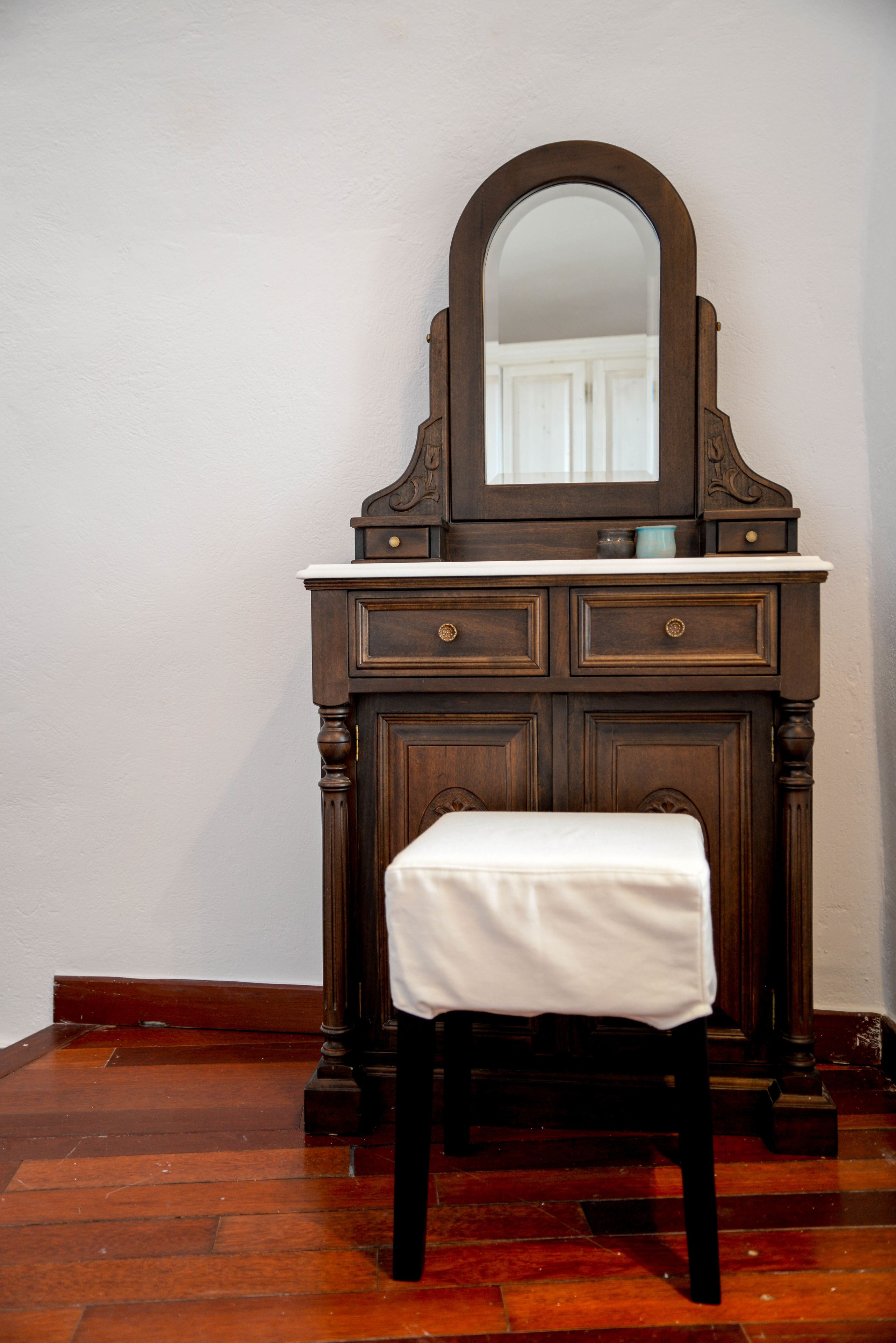 Un aparador de madera con espejo y cajones, un banco blanco adelante, y piso de madera.