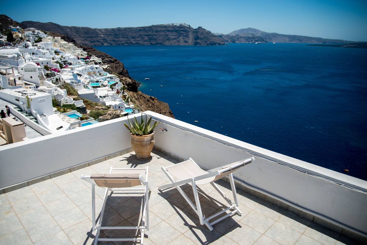 A terrace with white chairs and a potted plant, overlooking a coastal village with white buildings and a view of the sea.