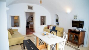 A living room with a sofa, a dining table, a bench, and a TV stand. - Genesis Cave Houses (Santorini)