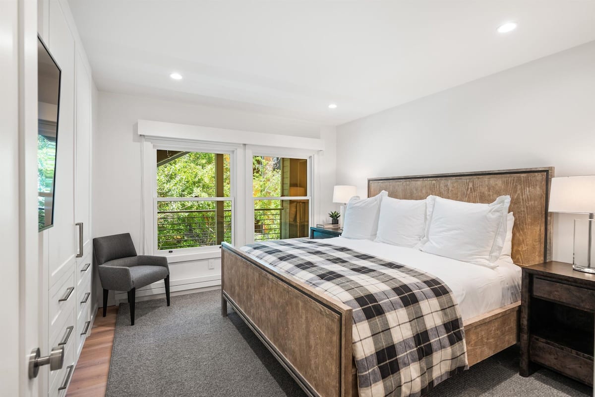 A bedroom with a large bed, a chair, a nightstand, and a window with a view of greenery.