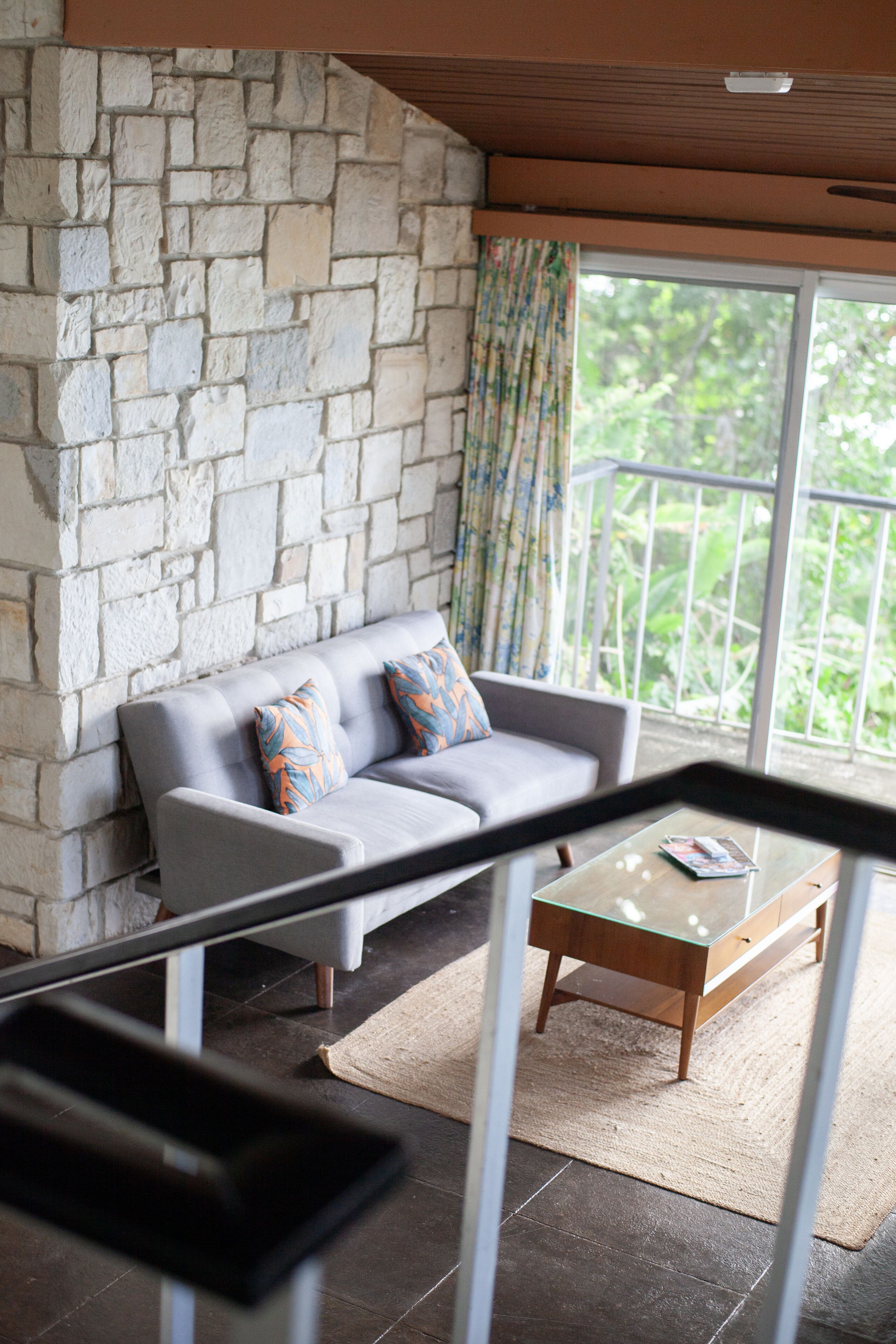 A living room with a stone wall, a grey sofa with patterned cushions, a glass coffee table, and a view of greenery through a balcony.