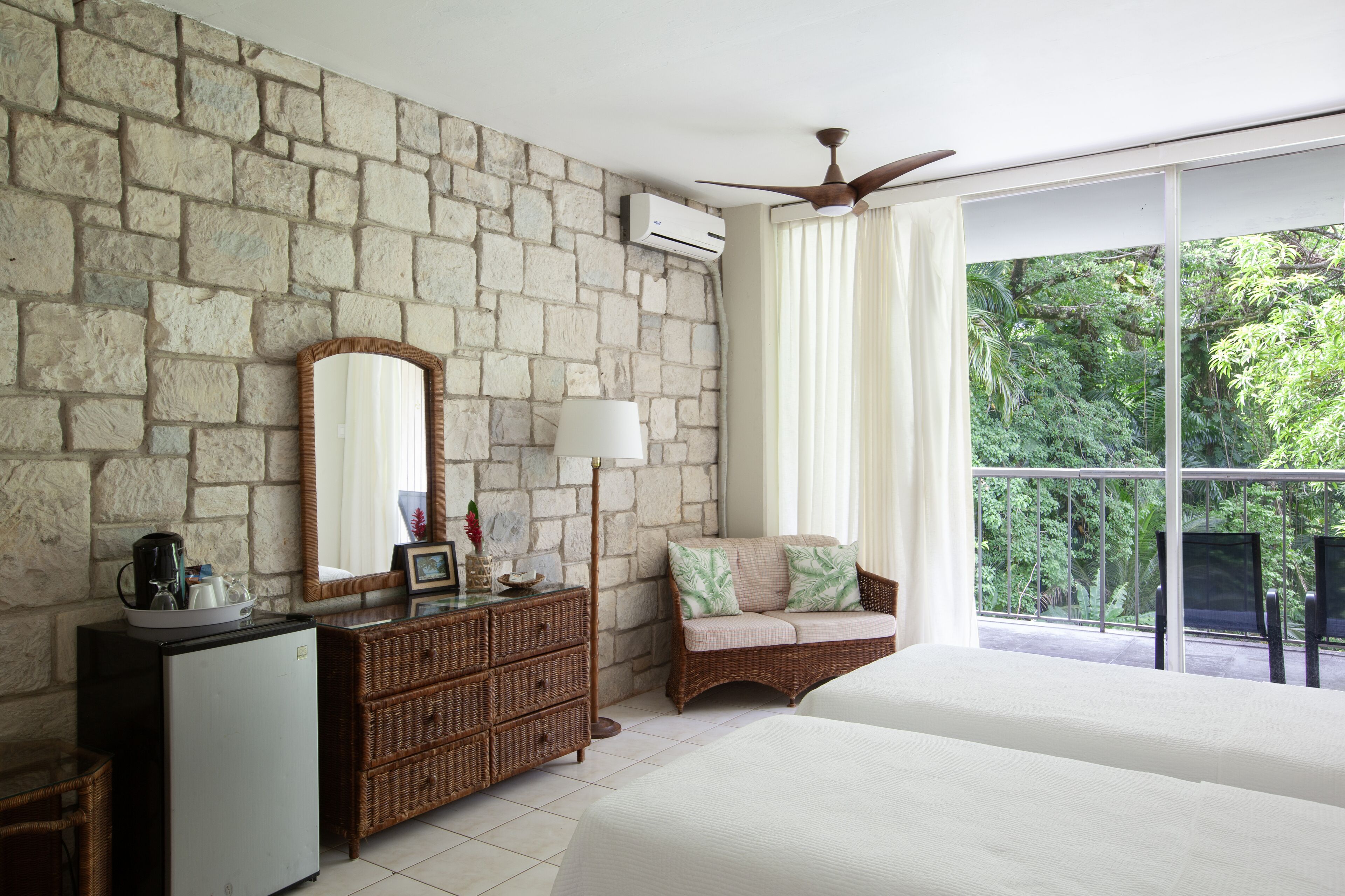 A bedroom with a stone wall, a ceiling fan, a large window with curtains, a wicker sofa, and a bed.