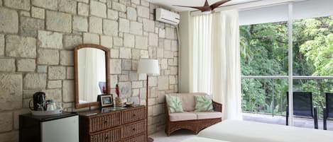A bedroom with a stone wall, a ceiling fan, a large window with curtains, a wicker sofa, and a bed.