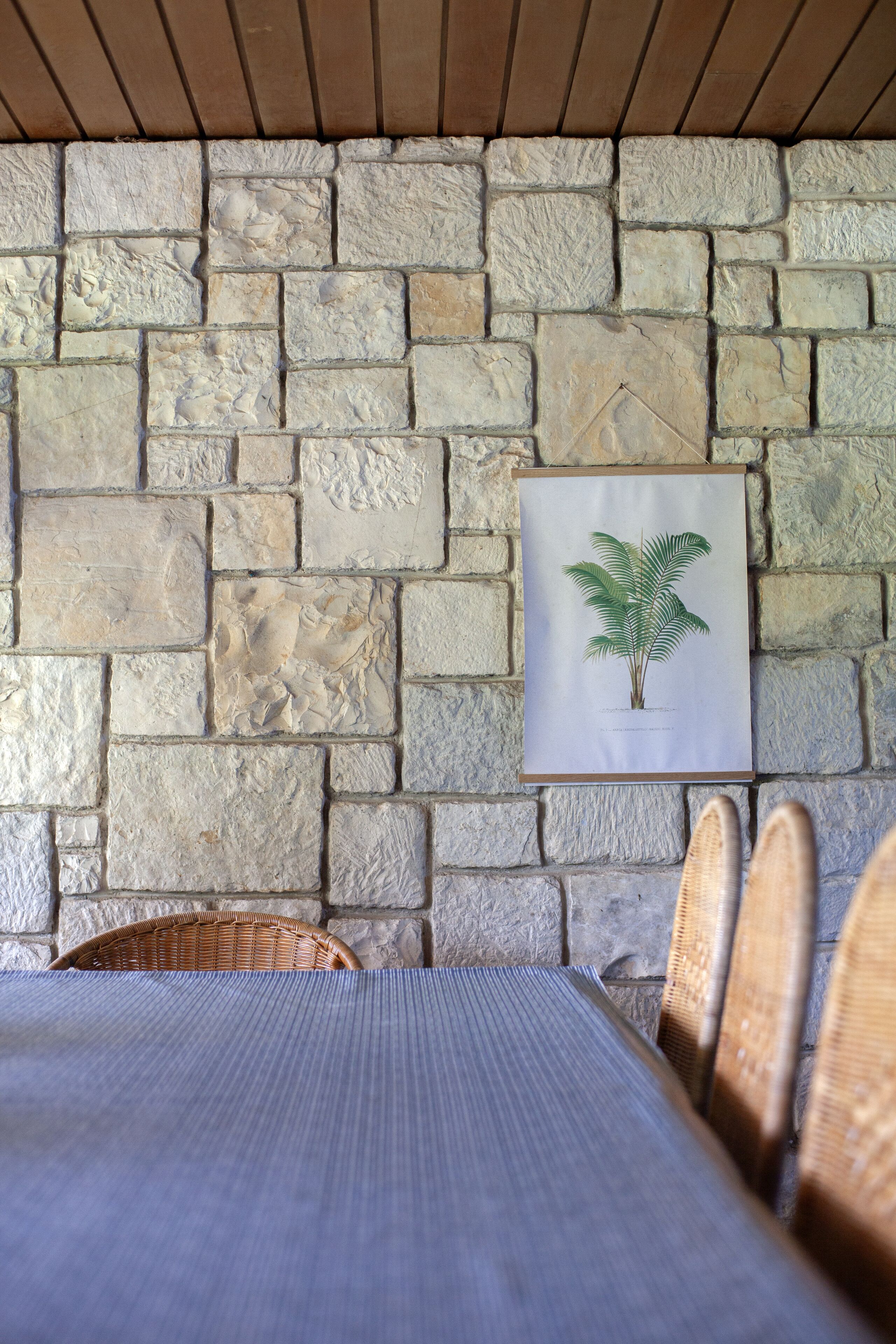 A stone wall with a framed botanical illustration, wicker chairs, and a striped tablecloth.
