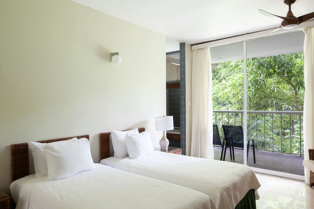 Ett hotellrum med två sängar, en balkong med utsikt över grönområden, en takfläkt och ett fönster med gardiner.