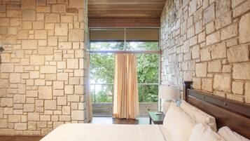 A bedroom with stone walls, a wooden ceiling, a large bed with white linens, a nightstand with a lamp, and a view of greenery through a window with curtains.