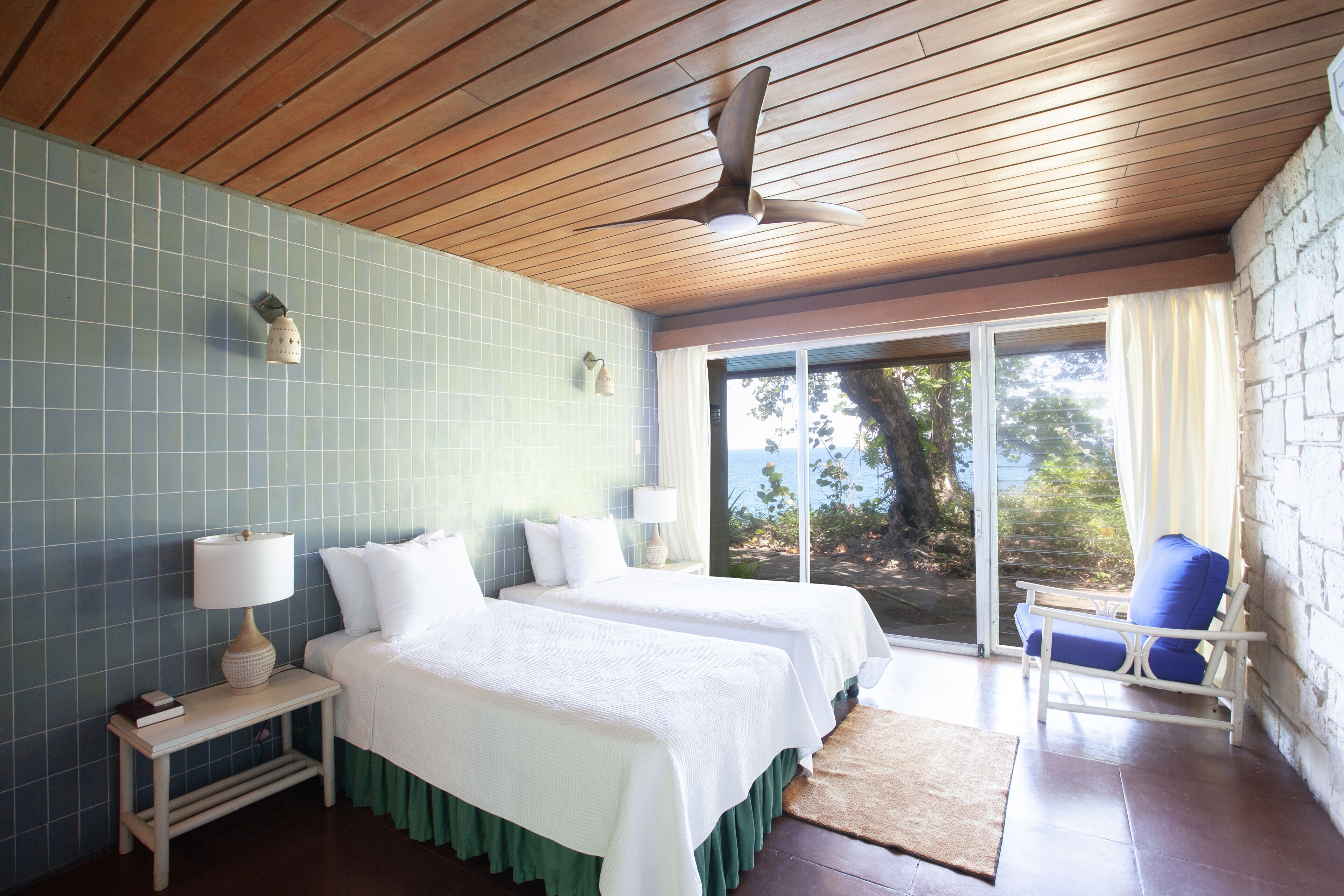 A room with two beds, a ceiling fan, a window with a view, and a chair.