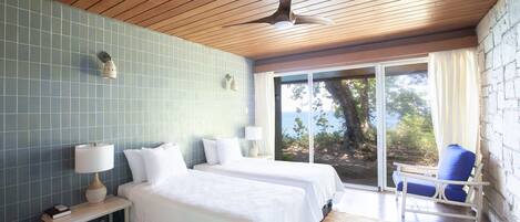 A room with two beds, a ceiling fan, a window with a view, and a chair.