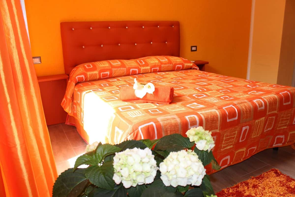A hotel room with a large bed, orange bedding, a nightstand, and a potted plant with white flowers.