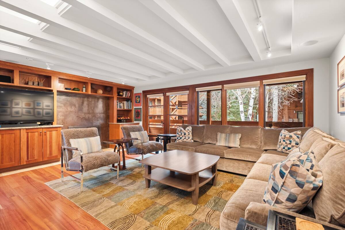 A spacious living room with a large wooden entertainment center, comfortable seating, and large windows offering a view of the outdoors.