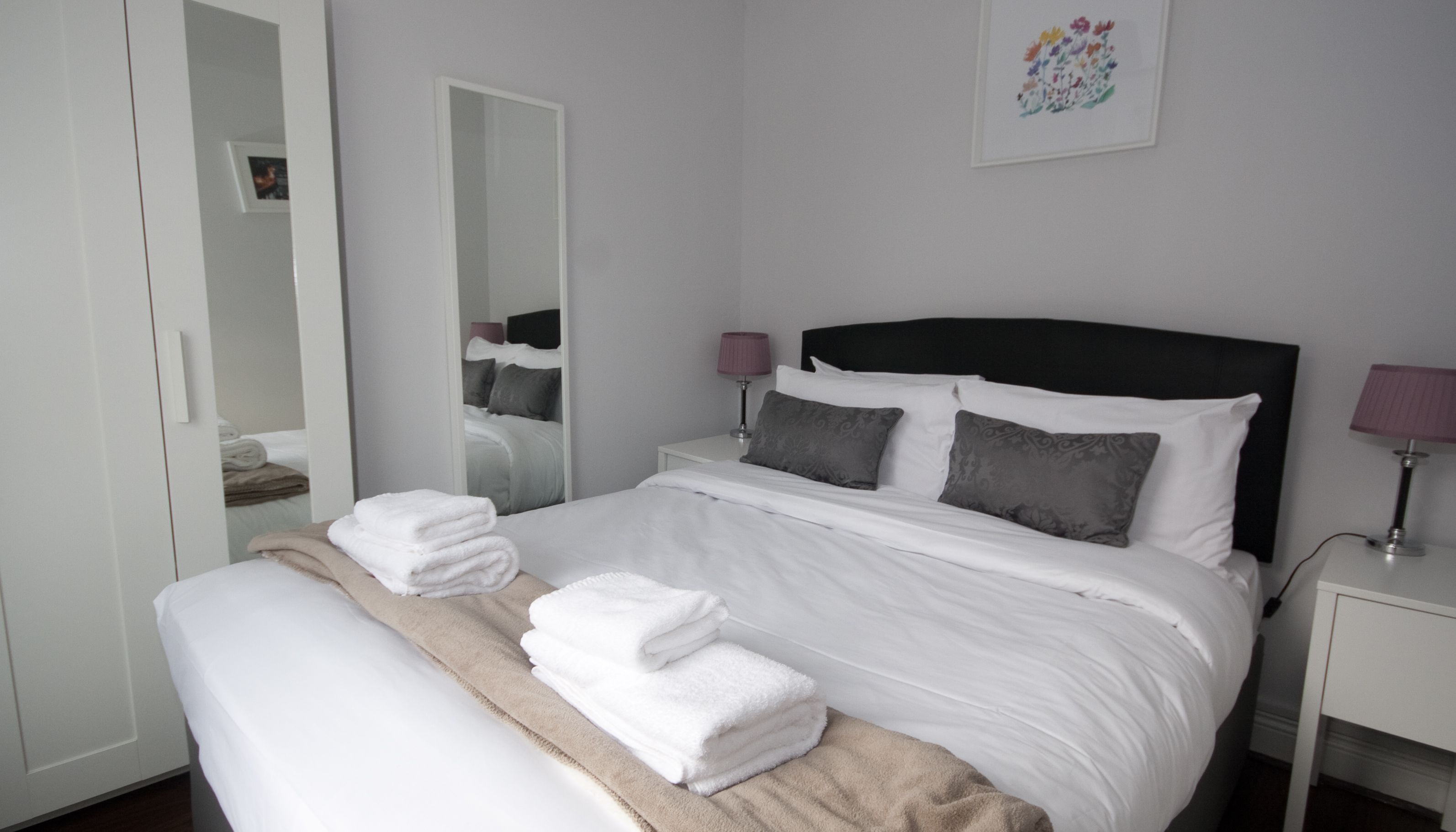Un lit bien fait, avec du linge de lit blanc et un plaid beige, une table de chevet avec une lampe et un miroir au mur.