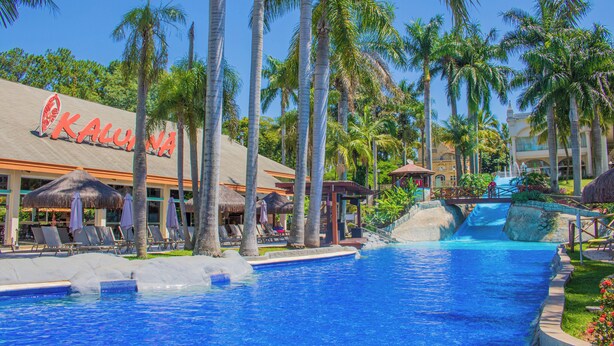 Ein Poolbereich einer Ferienanlage mit Palmen, Liegestühlen und einem Wasserfall.