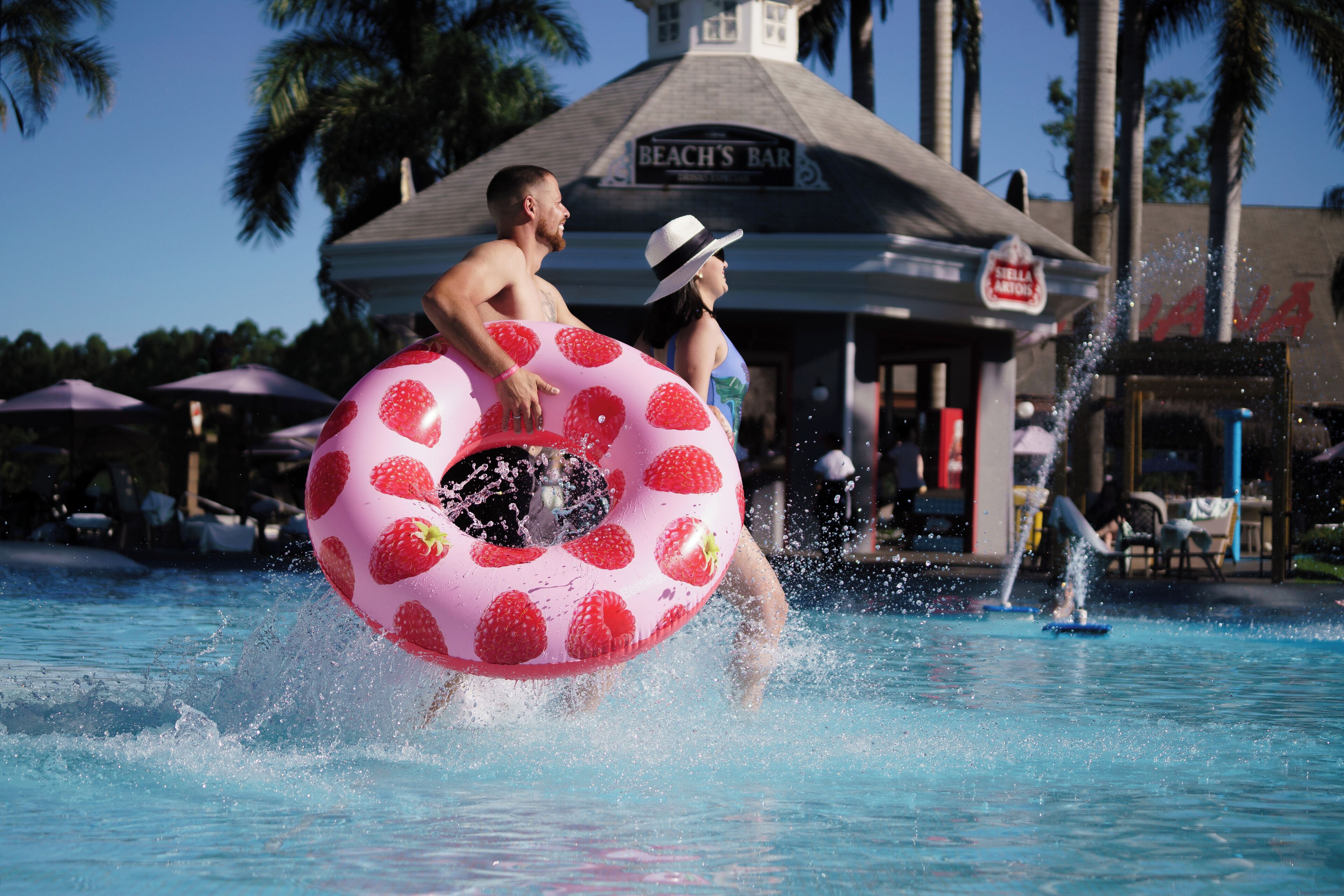 A man in a pool with a strawberry-patterned float, a beach bar, and palm trees in the background.