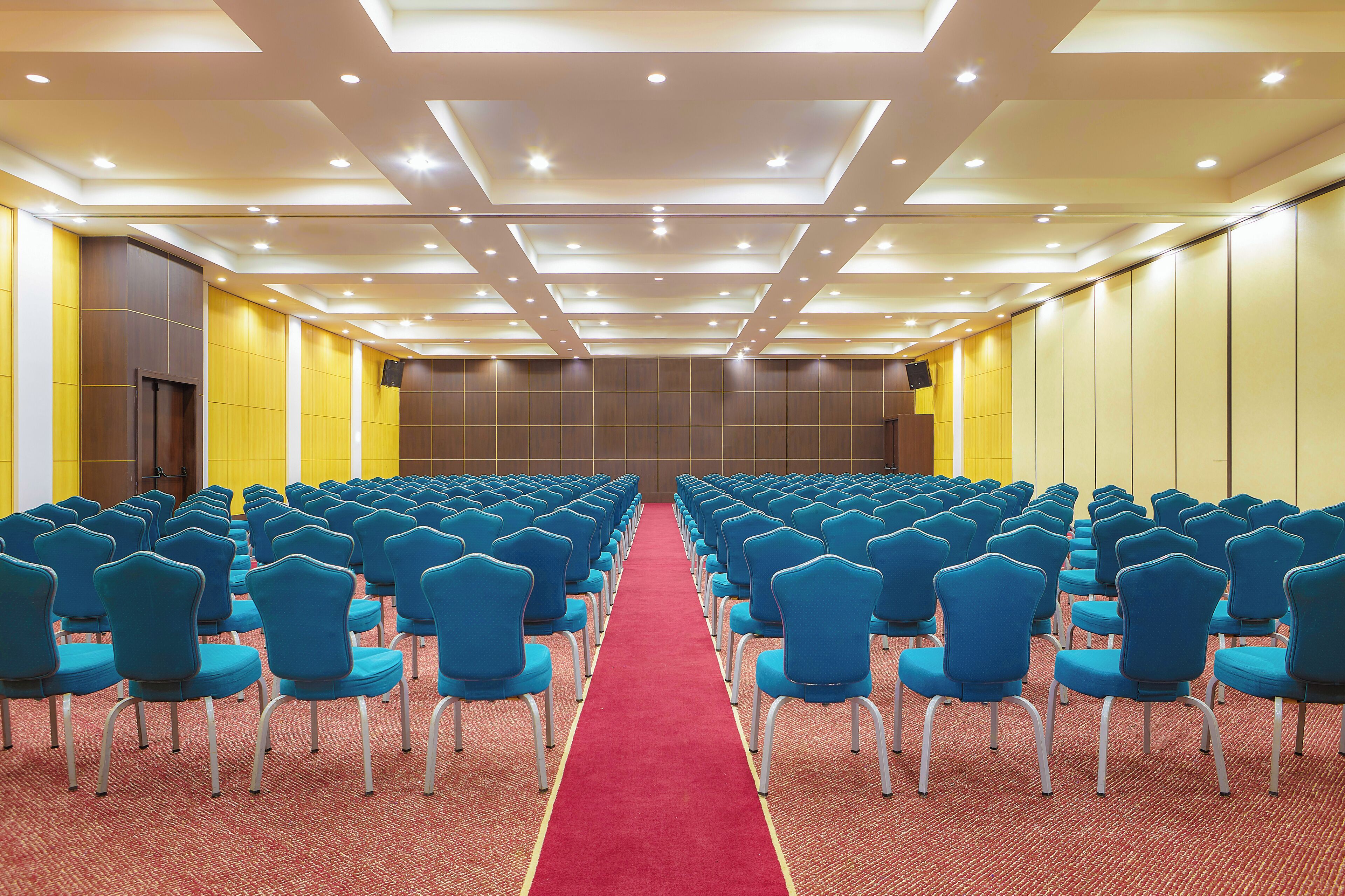 A conference room with rows of blue chairs, a red carpet, and recessed lighting on the ceiling.
