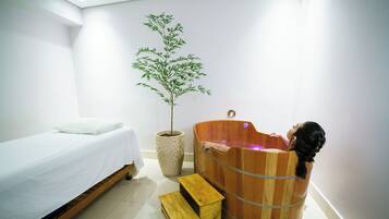 A room with a wooden bathtub, a bed with white bedding, and a potted plant.