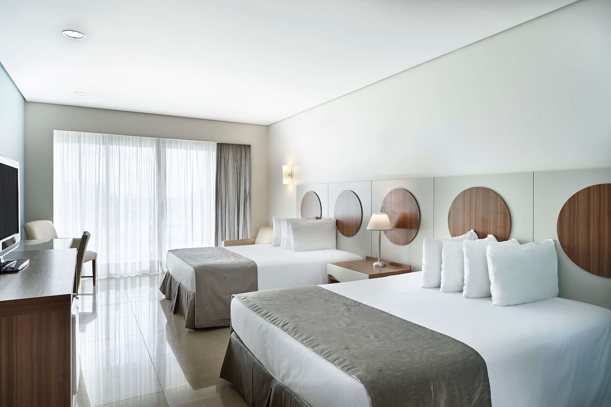Une chambre d’hôtel avec deux lits, un bureau avec une chaise, une télévision et une grande fenêtre avec des rideaux.