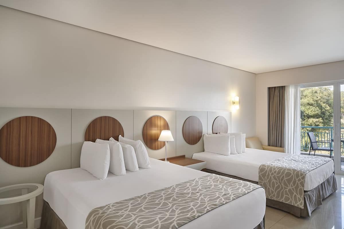 Une chambre d’hôtel avec deux lits, une horloge murale ronde, une table de chevet, une lampe et un balcon donnant sur la verdure.