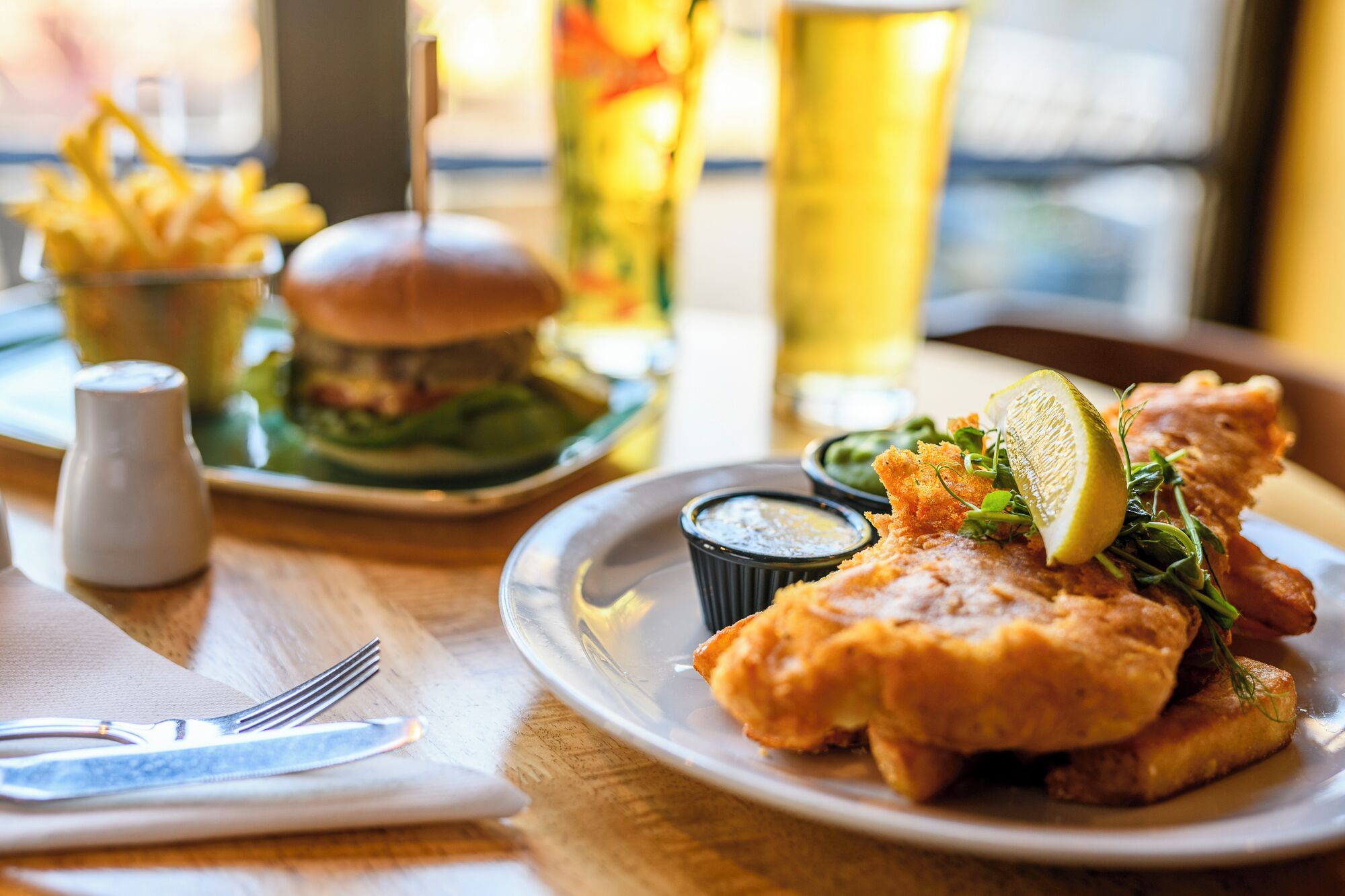 Un plato de pescado con patatas fritas acompañado de salsa tártara, una hamburguesa y una cerveza en una mesa.