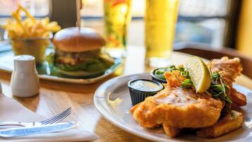 Un plato de pescado con patatas fritas acompañado de salsa tártara, una hamburguesa y una cerveza en una mesa.