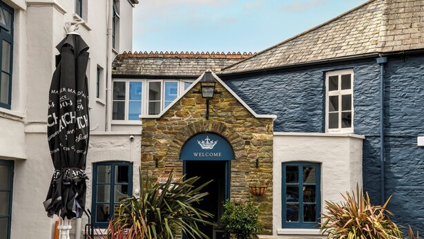 A building with a stone archway entrance, a 'WELCOME' sign above the door, and a flag with text hanging on the left. - The Crown Hotel (Lynton)