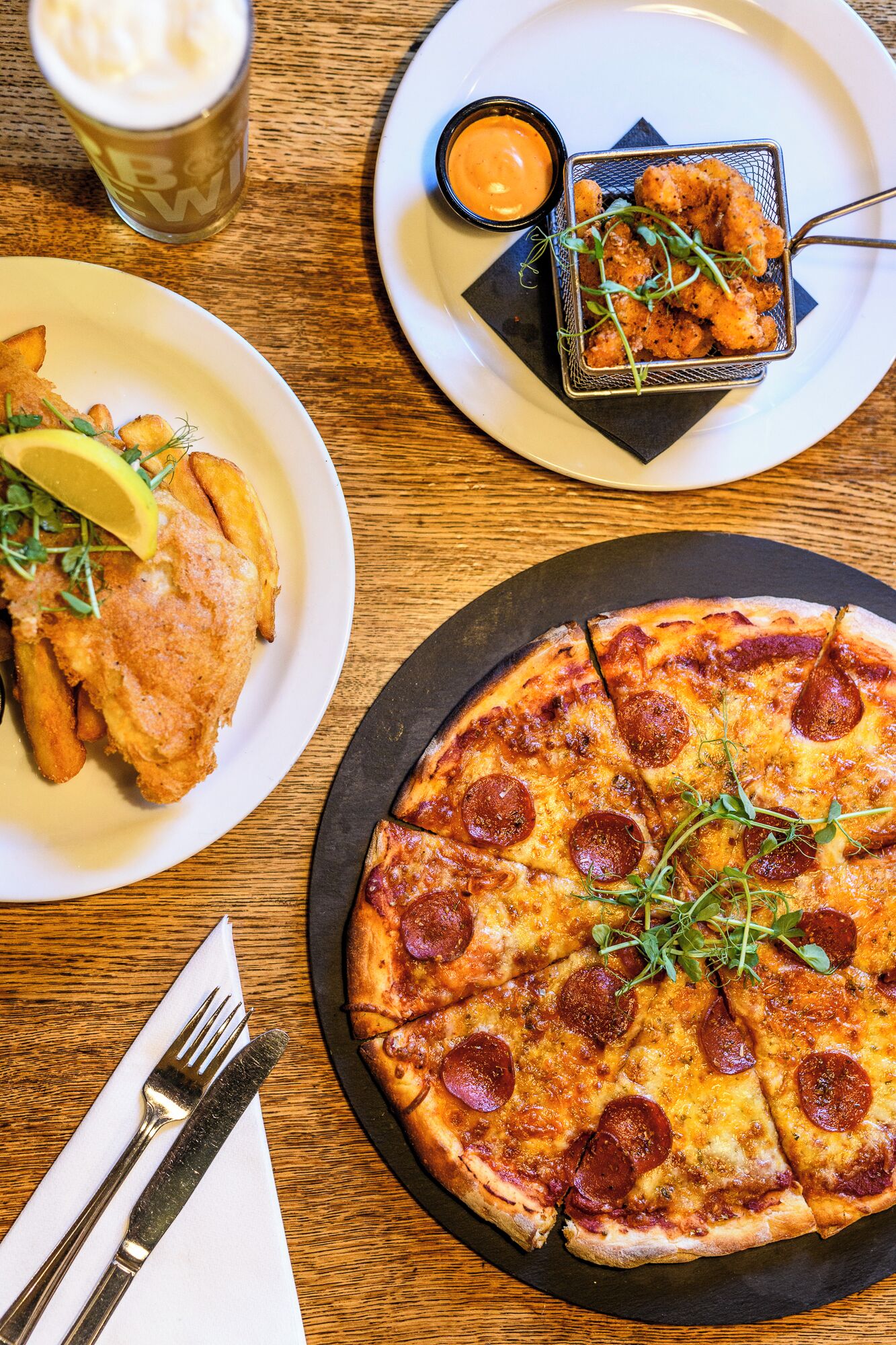 A pizza with pepperoni slices, a plate of fried fish, and a small bowl of sauce on a wooden table.