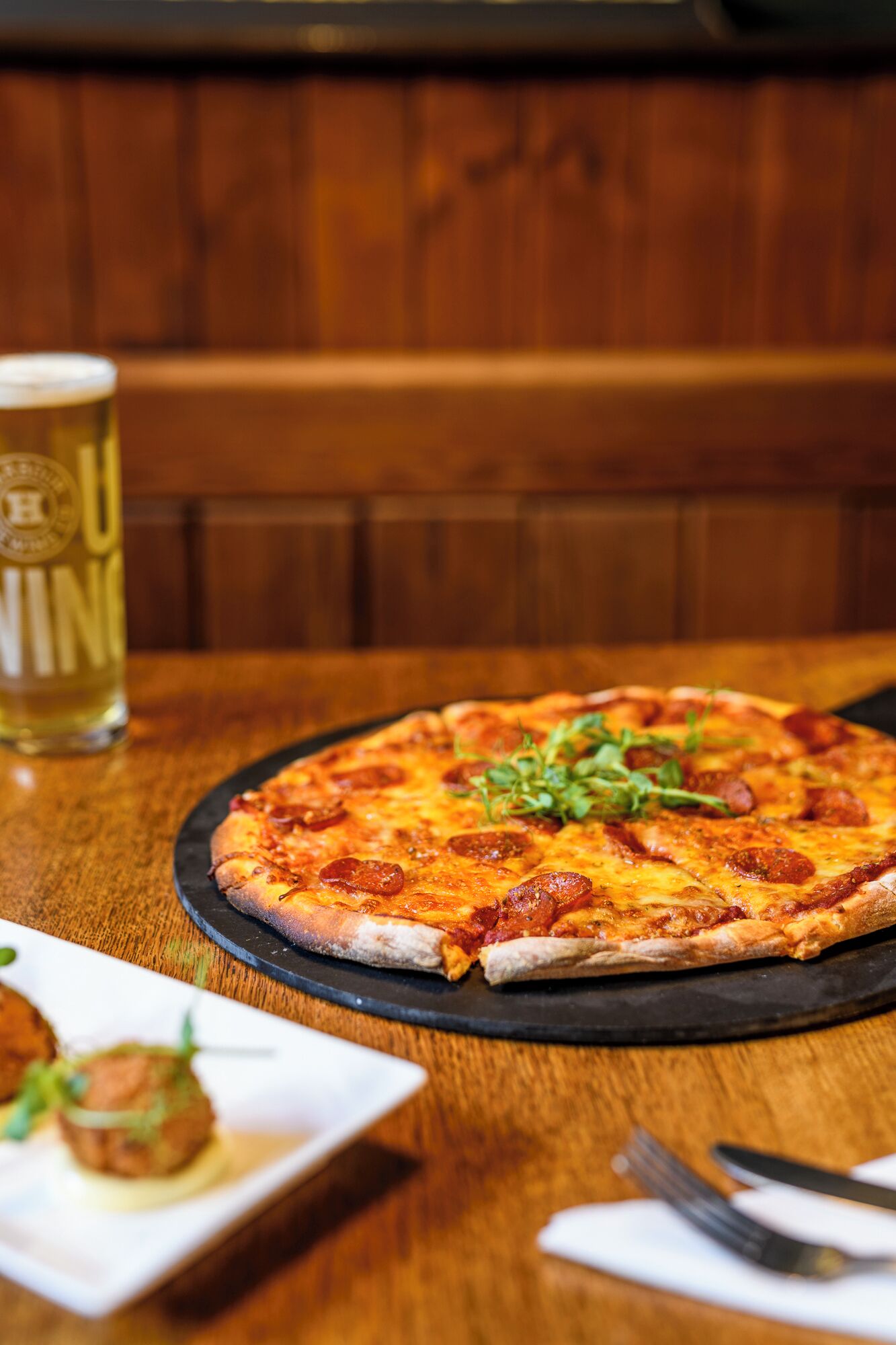 Una pizza sobre un plato negro, con un vaso de cerveza y un tenedor sobre una mesa de madera.