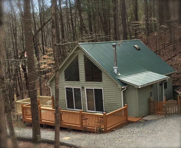 A cabin with a green roof, wooden deck, and a gravel driveway.