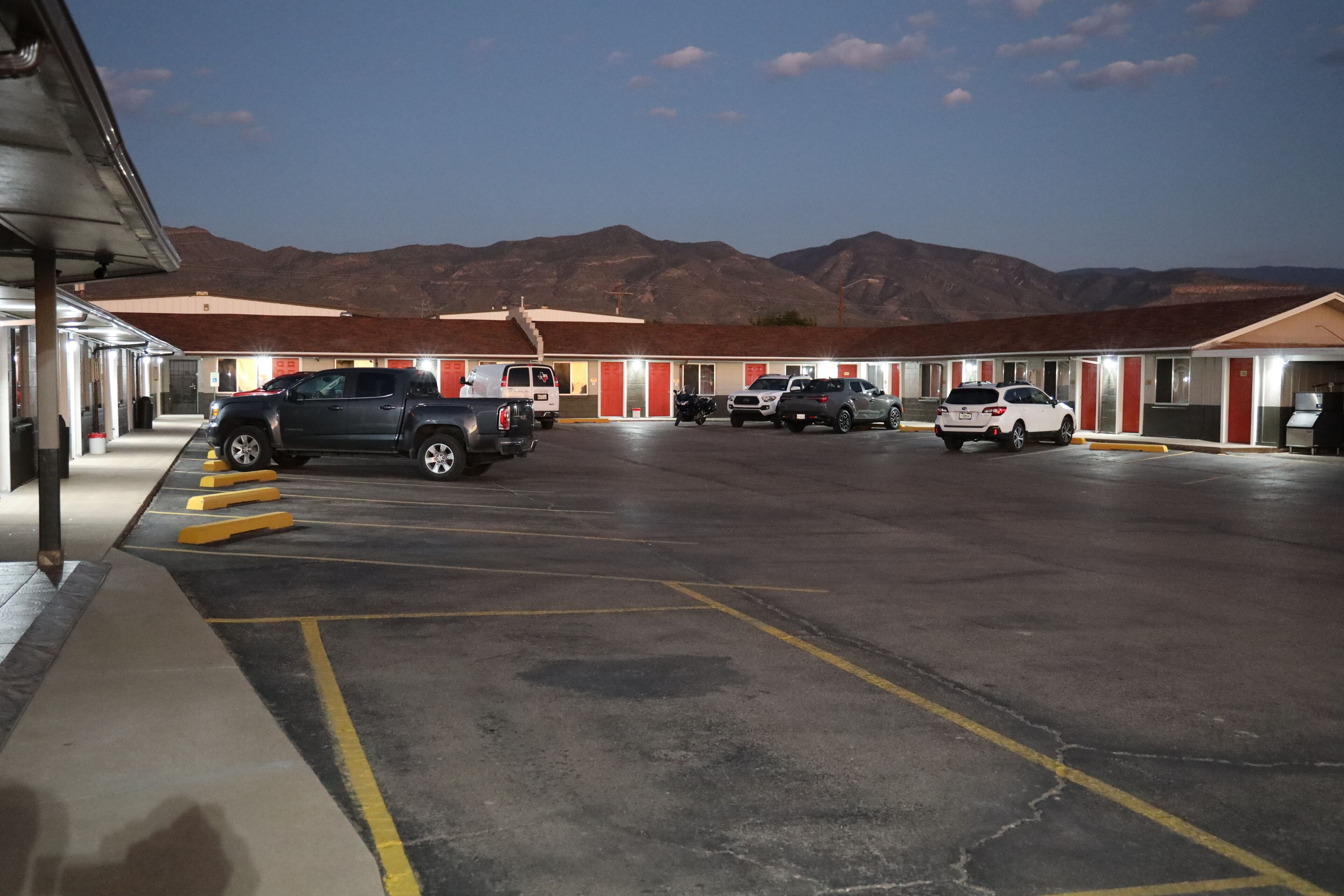 A motel with multiple rooms, a parking lot, and mountains in the background.