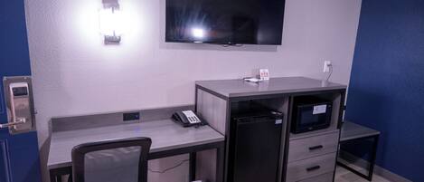 A hotel room with a desk, chair, television, microwave, and a trash bin.