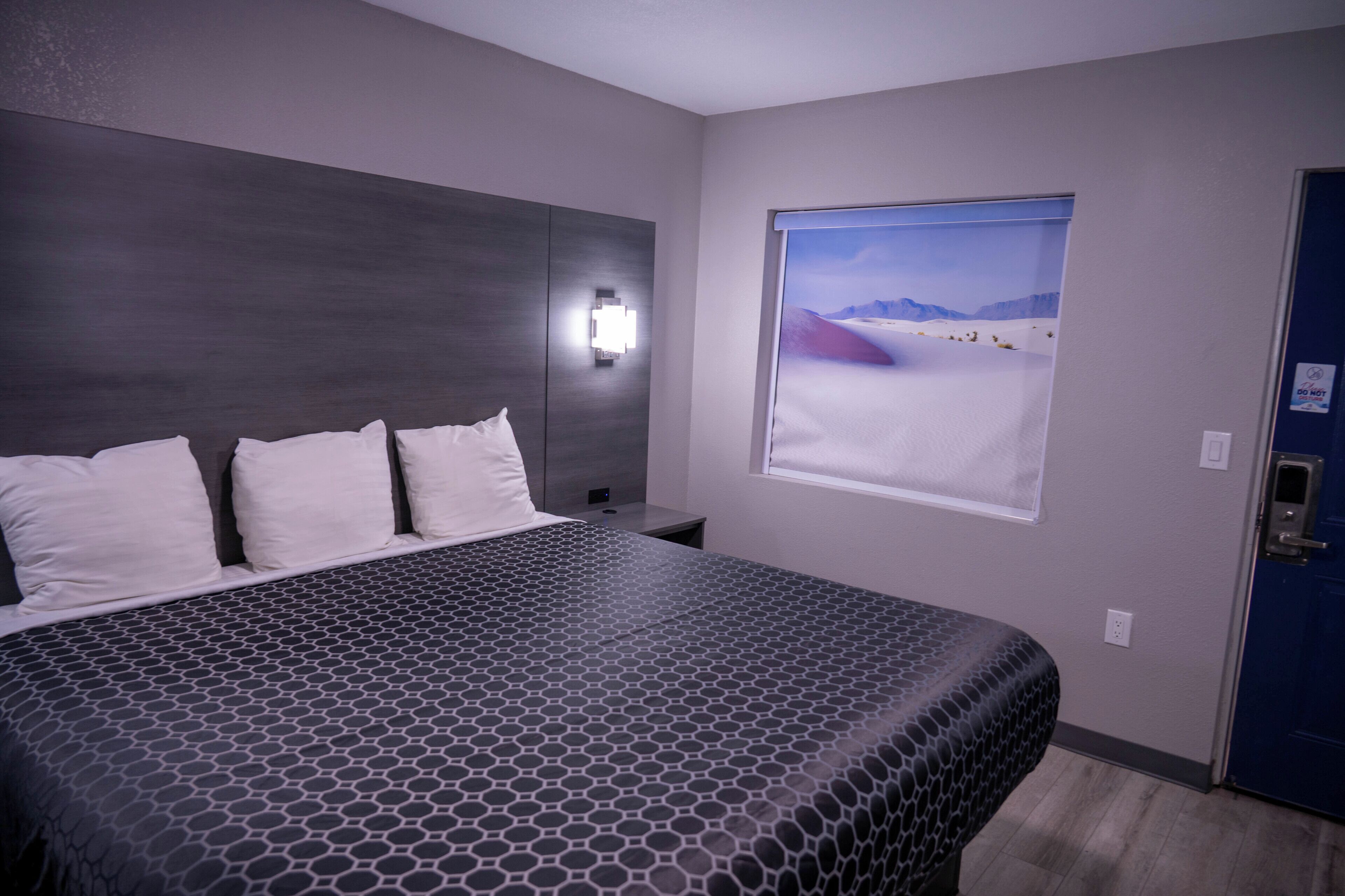 Hotelkamer met een groot bed, nachtkastje, wandlamp en raam met uitzicht op een woestijn.