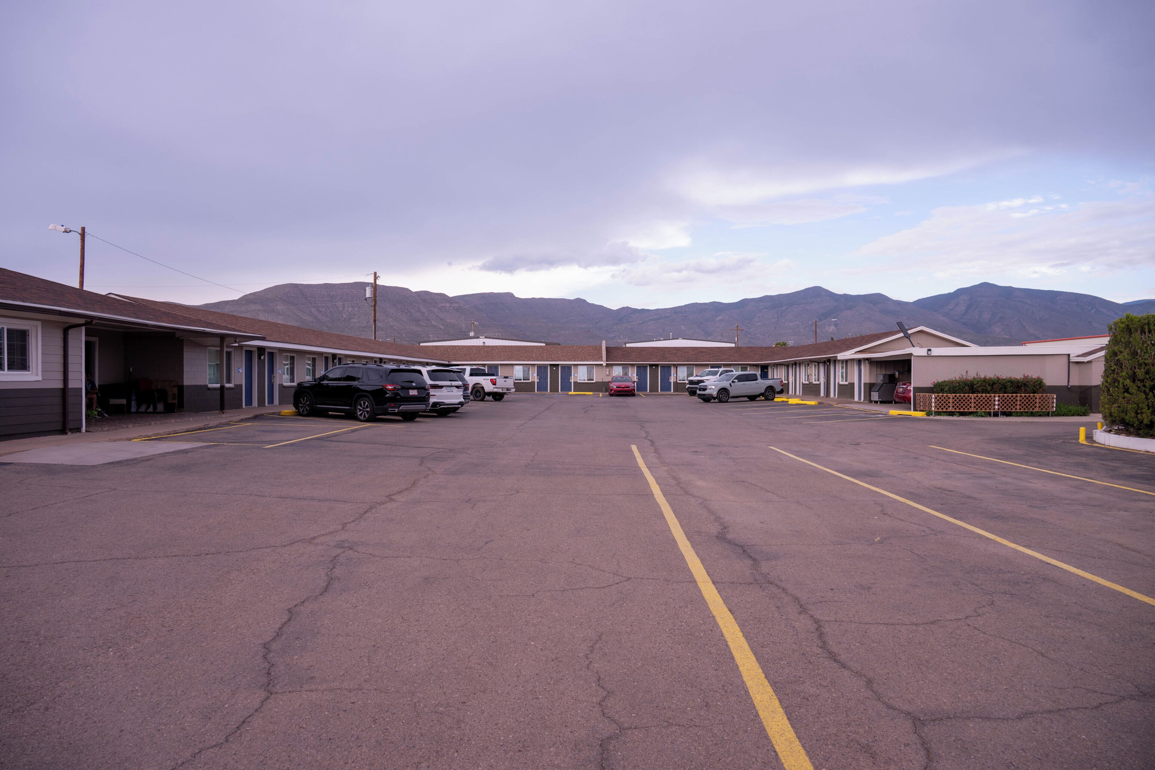A parking lot with several cars parked in front of a row of motel-style buildings with mountain views in the background.