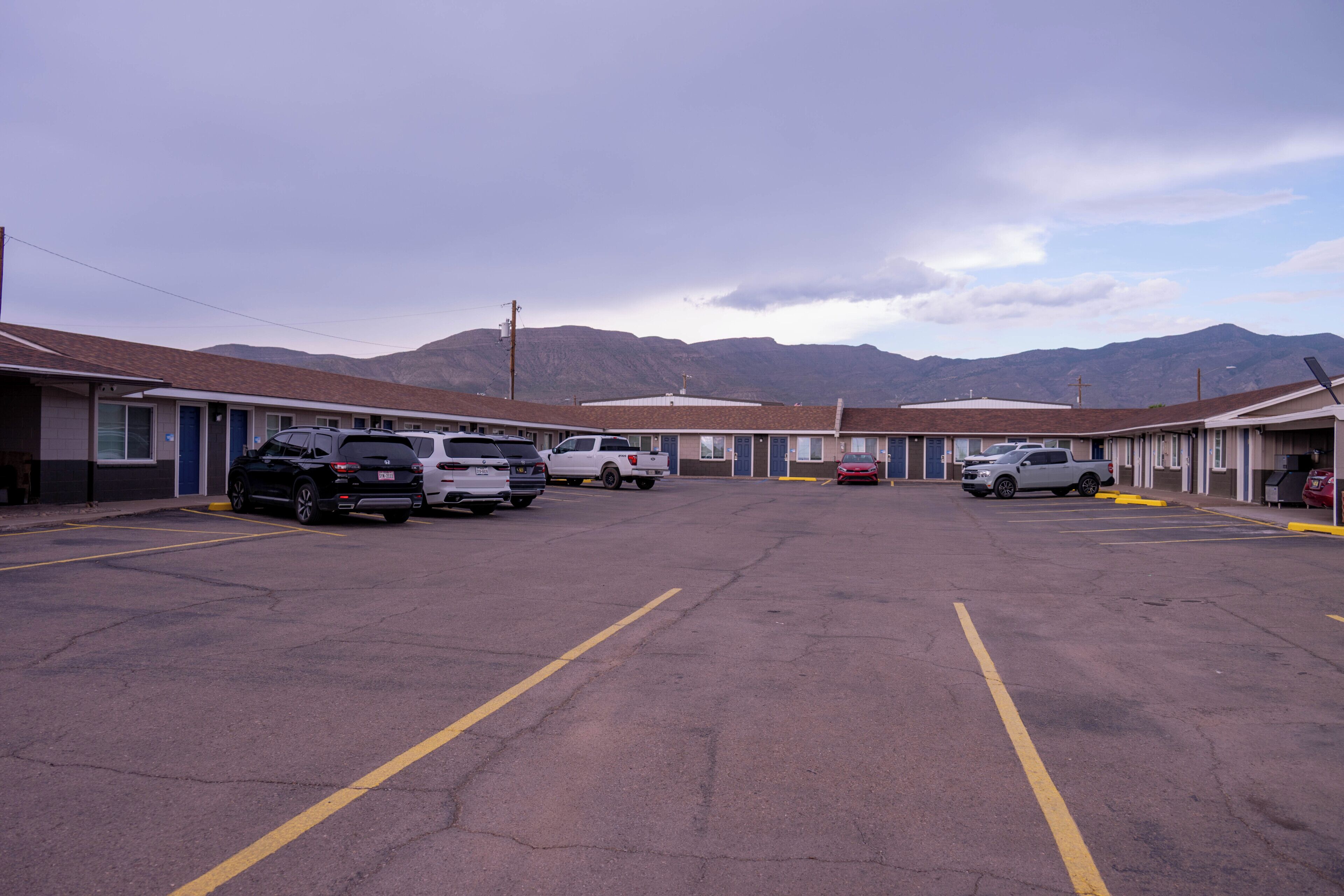 A parking lot with several vehicles, including SUVs and a pickup truck, in front of a row of motel buildings with a mountainous backdrop.