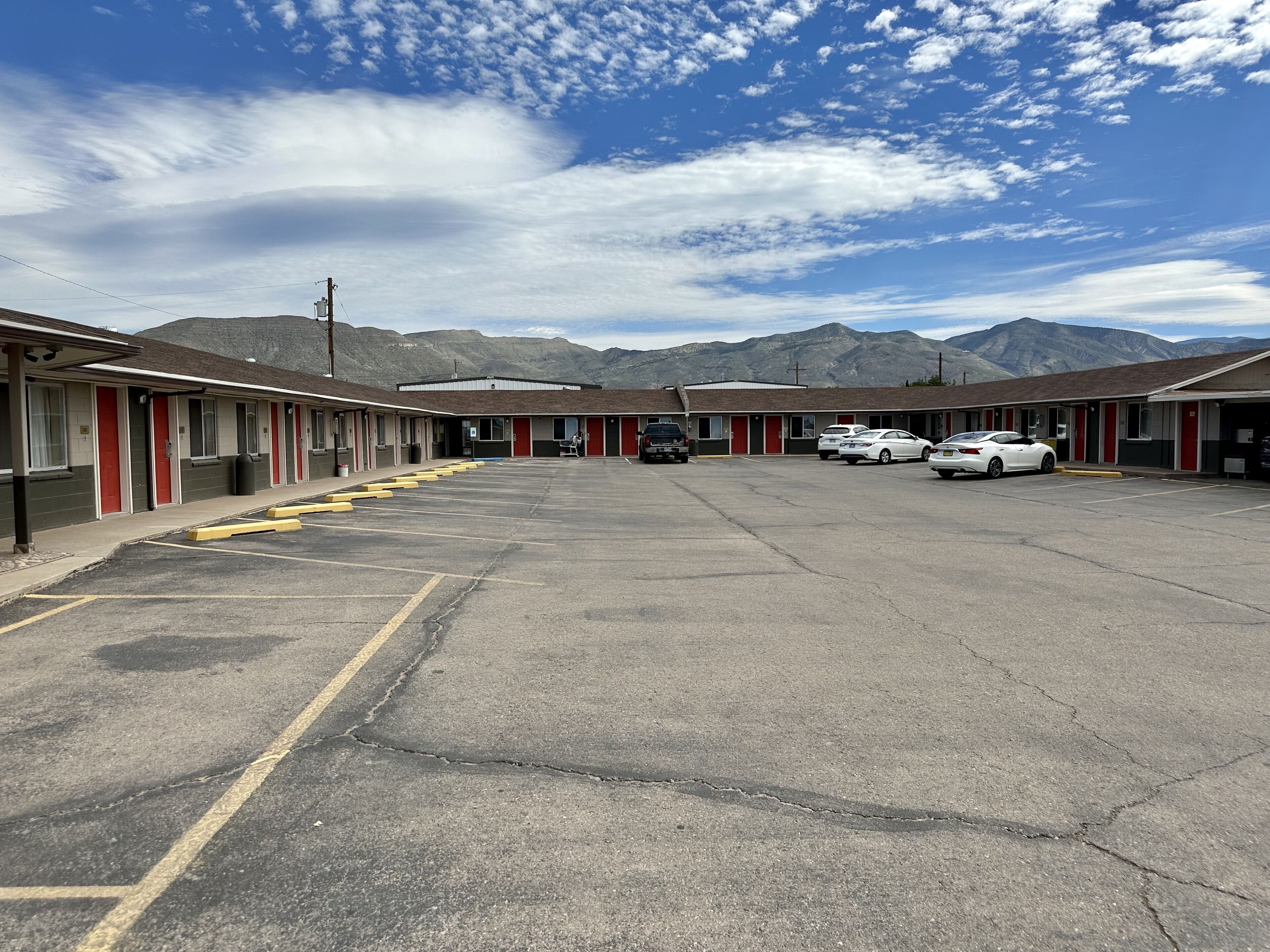 A motel with a parking lot, several cars parked, and mountains in the background.