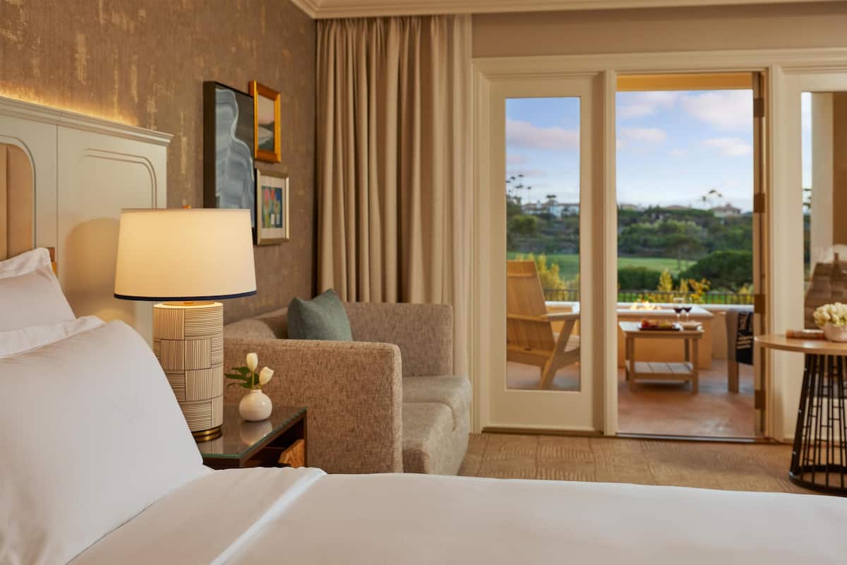 Habitación de hotel con cama, lámpara, silla, mesita y vistas al exterior por una puerta corredera.