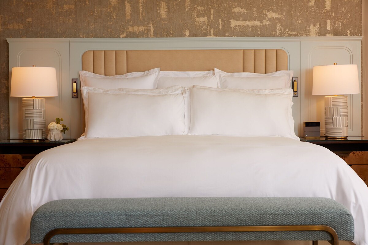 Una cama bien hecha con sábanas blancas, con dos mesitas de noche con lámparas y un jarrón de flores.