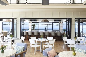 A spacious indoor dining area with multiple tables and chairs, large windows offering a view of the sea, and a bar area with high stools.