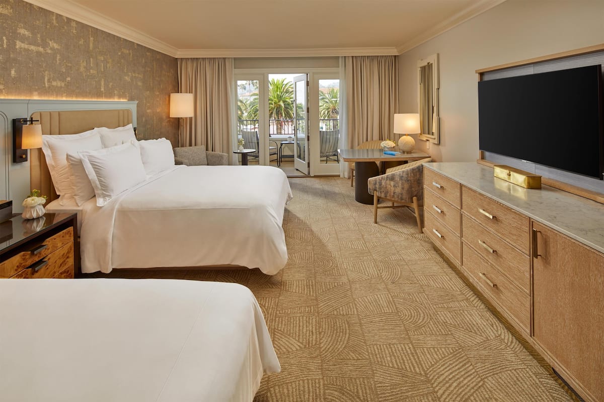Habitación de hotel con dos camas, televisión, escritorio con silla y balcón con vistas a palmeras.