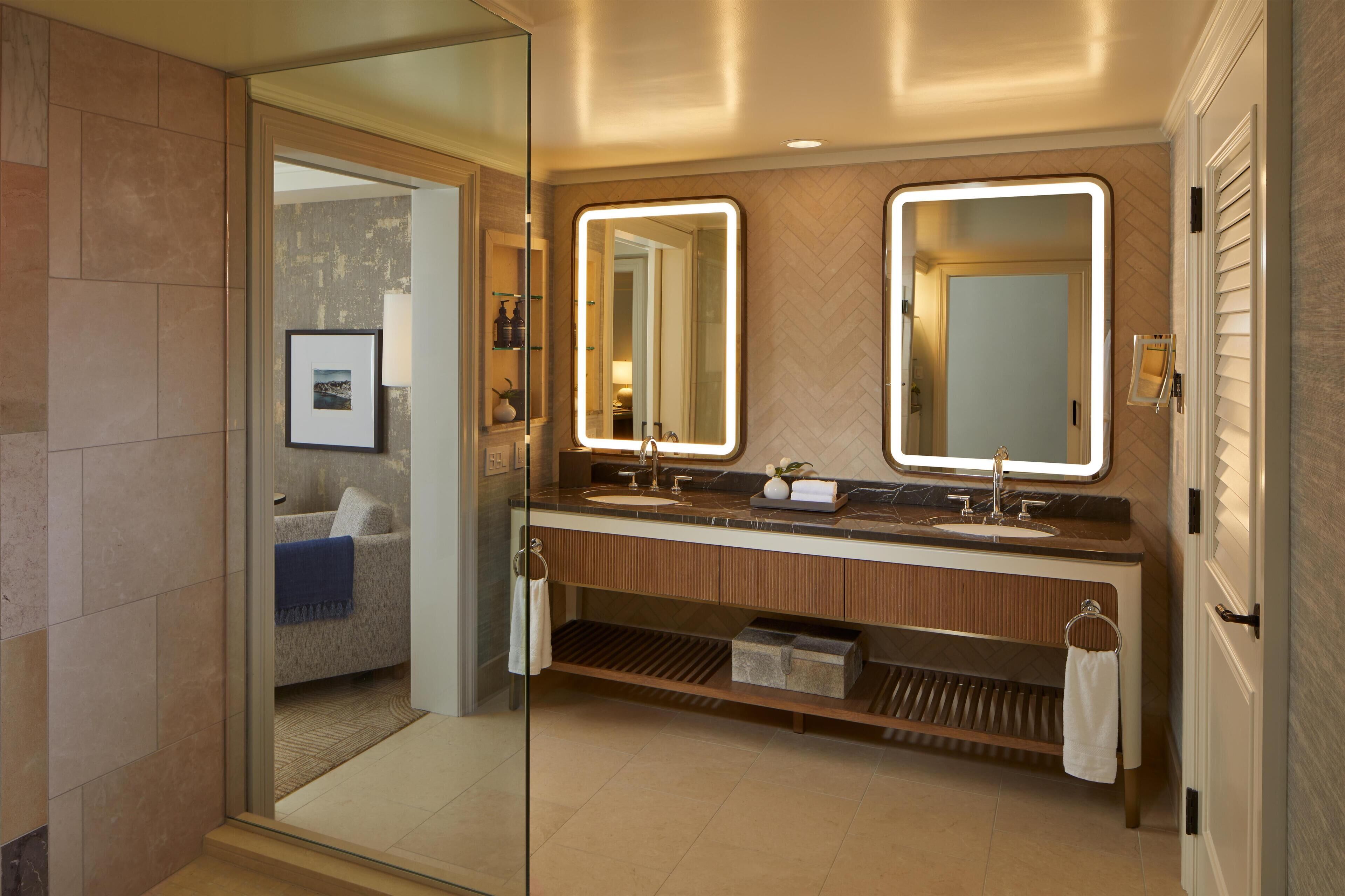 Un baño moderno con cabina de ducha de vidrio, lavamanos doble con espejos iluminados y vista hacia un dormitorio con cama y un cuadro en la pared.