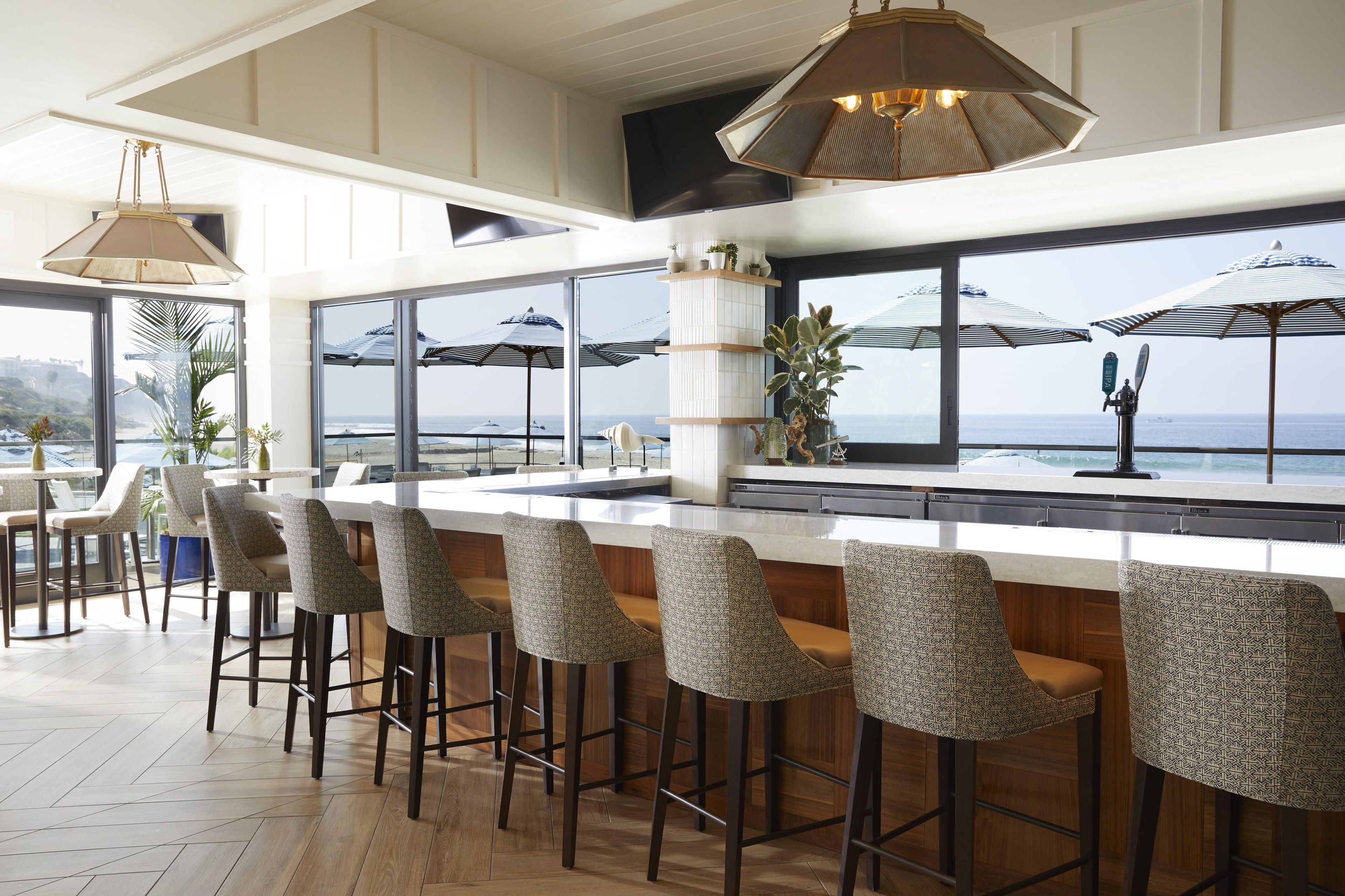 Zona de bar con taburetes altos, una encimera blanca y ventanas amplias con vistas al mar.